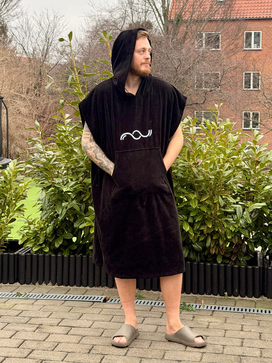 Person wearing a black sleeveless poncho with a logo, standing outdoors on a paved area with greenery and buildings in the background.