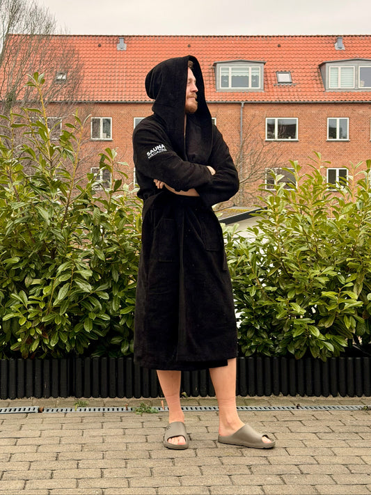 Person wearing a black robe with a hood in an outdoor setting with greenery and a building in the background.
